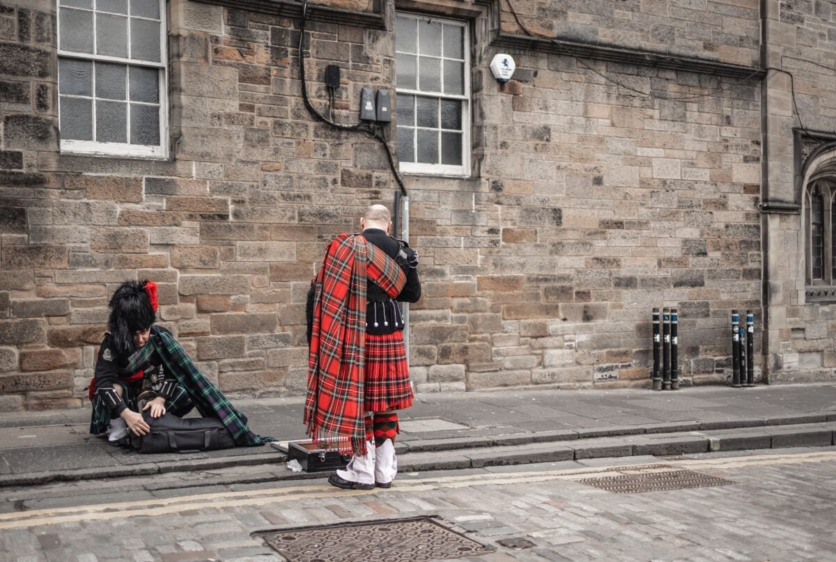 Two bagpipers in traditional Scottish kilts prepare instruments on a cobblestone street.