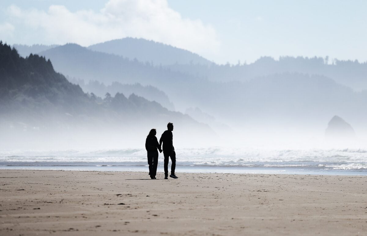 pexels 34260821 | Two Scots Abroad Romantic couple holding hands walking along Cannon Beach with coastal backdrop.