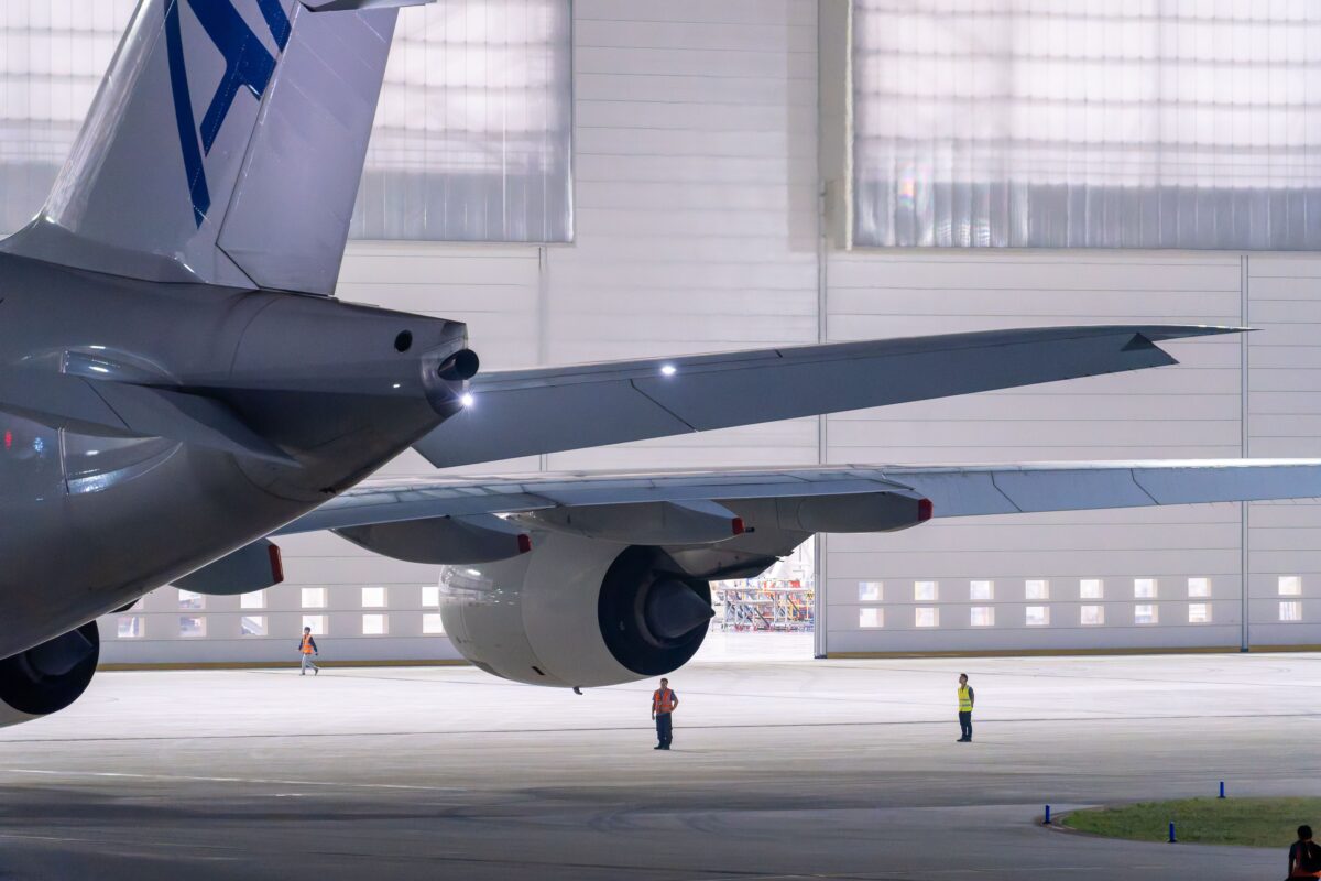 Large commercial airplane parked in an illuminated airport hangar with maintenance staff visible.