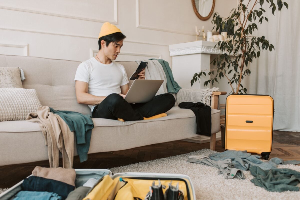 Adult male traveler planning trip at home with laptop and smartphone, surrounded by luggage.