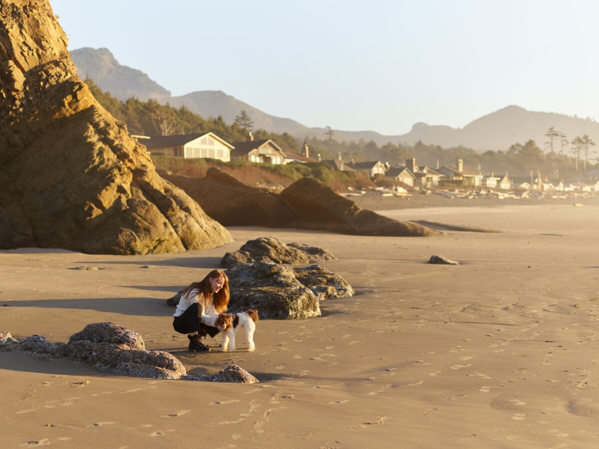 pexels 7978073 | Two Scots Abroad A woman pets her dog on a picturesque beach with rocks and a village in the background during sunset.