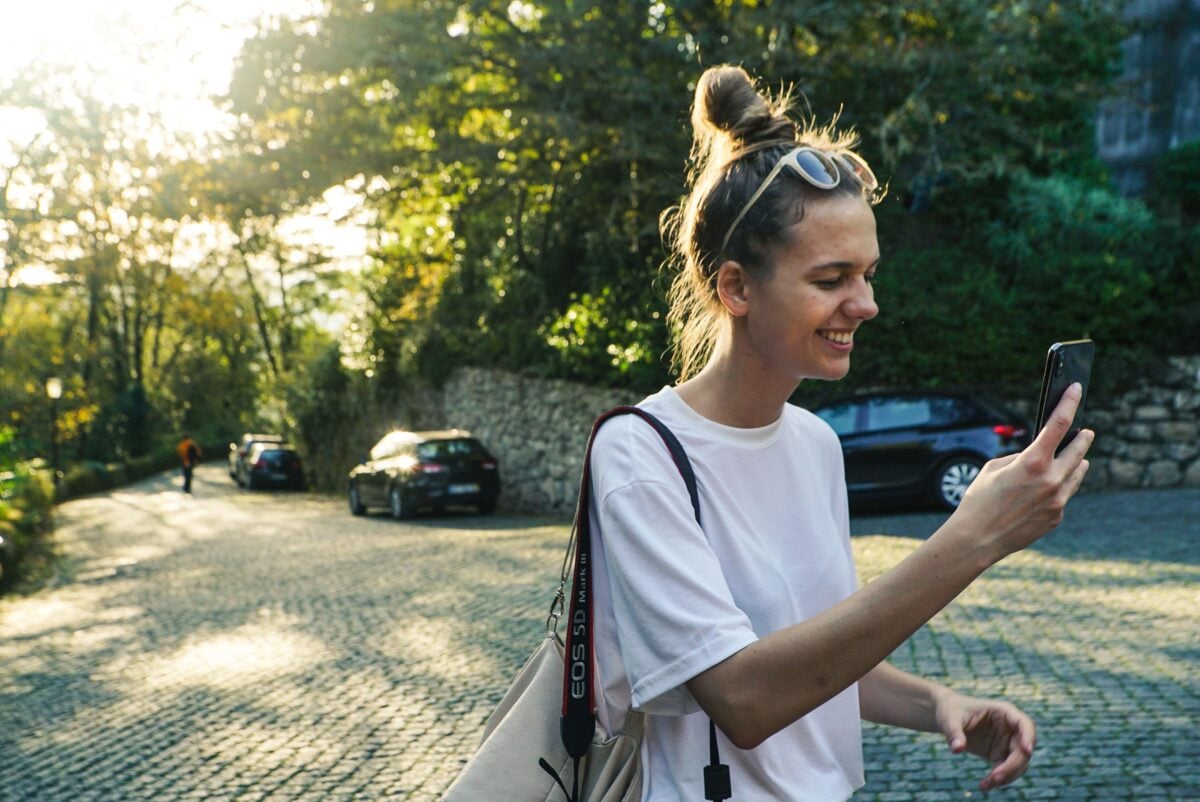 pexels 1551126 | Two Scots Abroad A joyful woman smiles as she checks her phone on a sunlit cobblestone street with greenery.