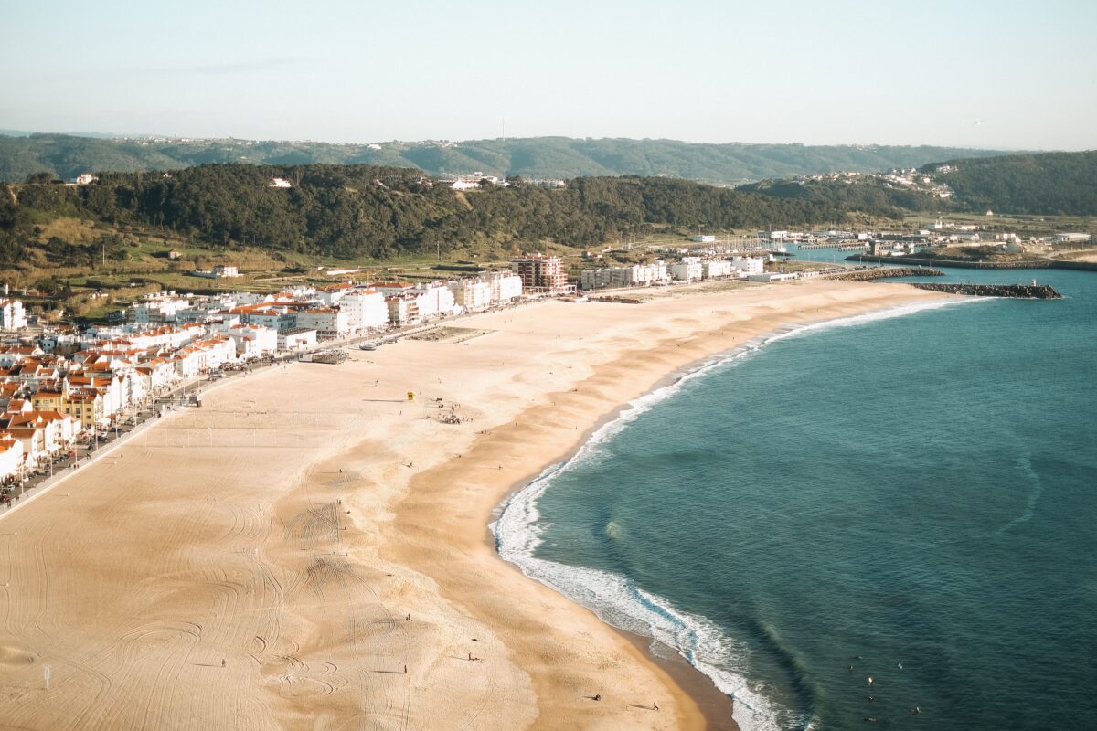 Aerial photography of Nazare beach with golden sands and vivid blue ocean against distant hills.