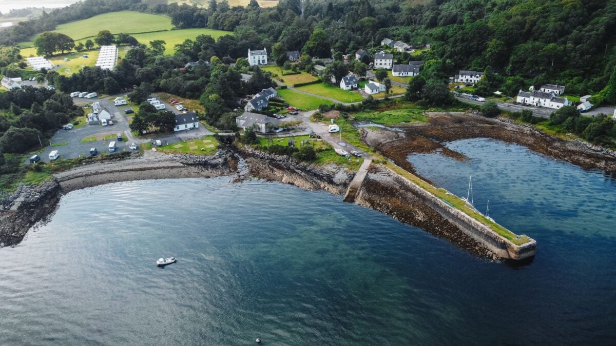 Aerial view of a picturesque coastal village in Scotland on a clear day, lush greenery and water visible.