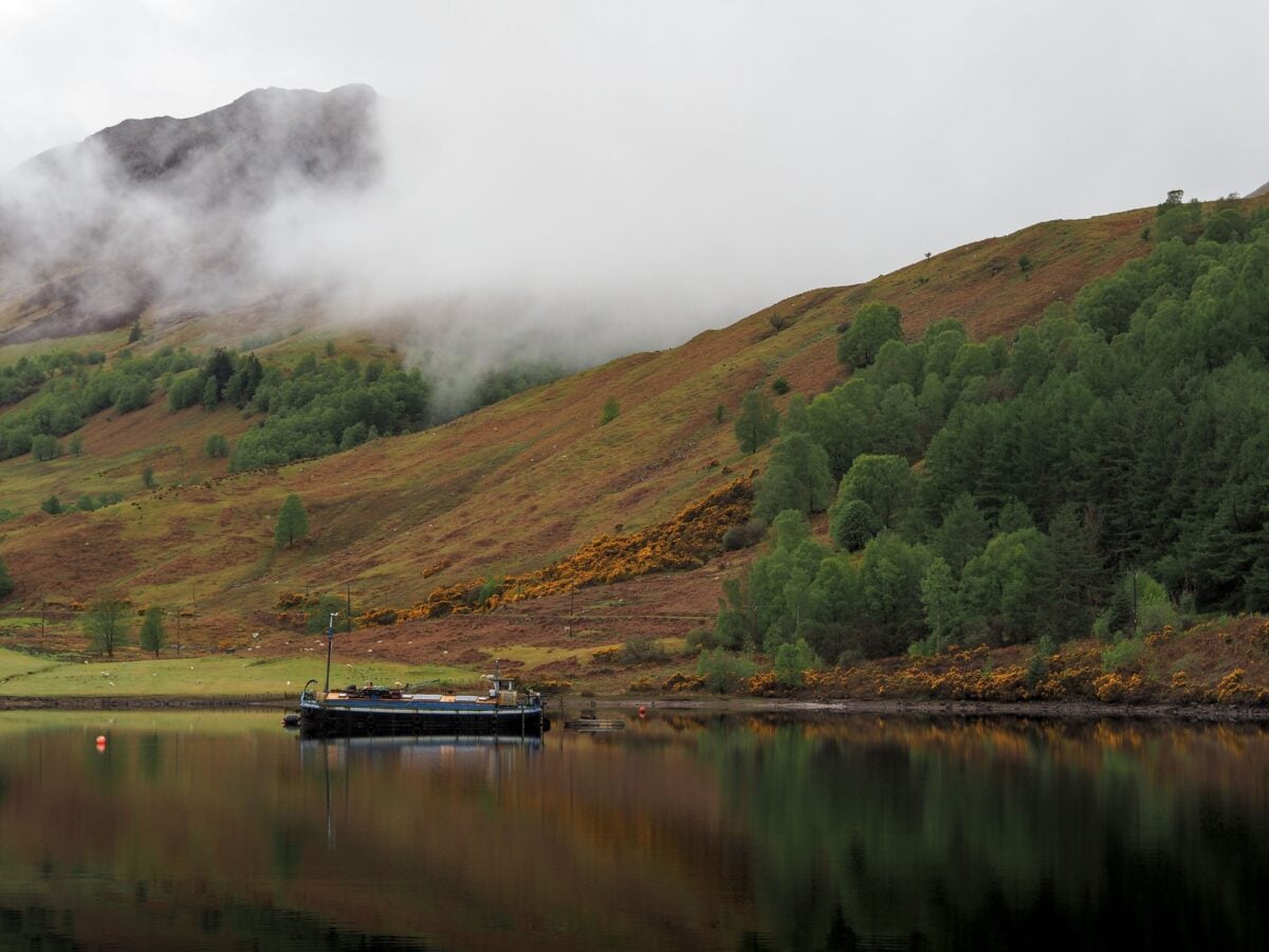 pexels 37591043 | Two Scots Abroad A serene morning at a Scottish loch with misty mountains and a tranquil boat reflecting on still waters.