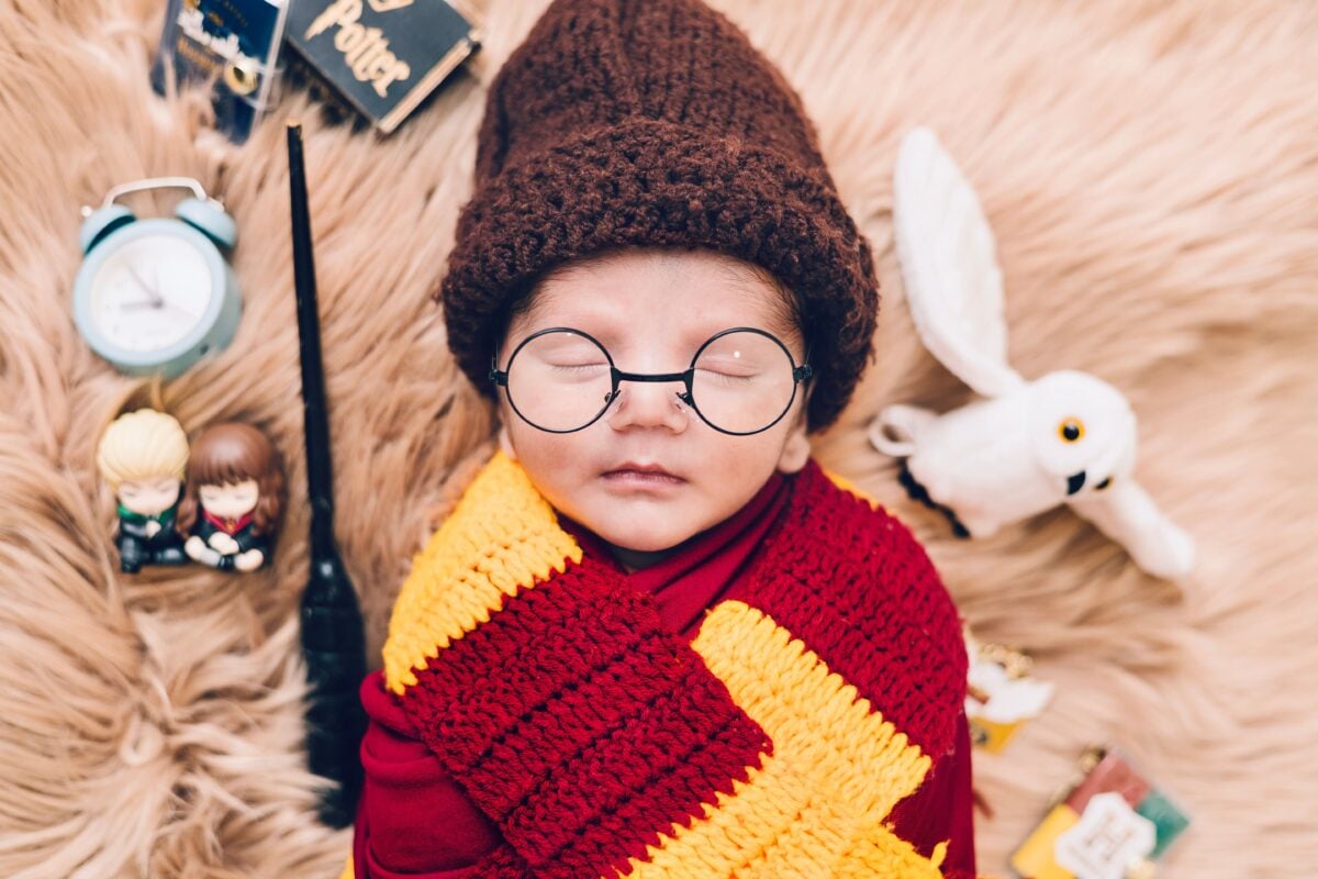 A cute newborn baby dressed in a wizard costume with glasses and a scarf, lying on a soft blanket.