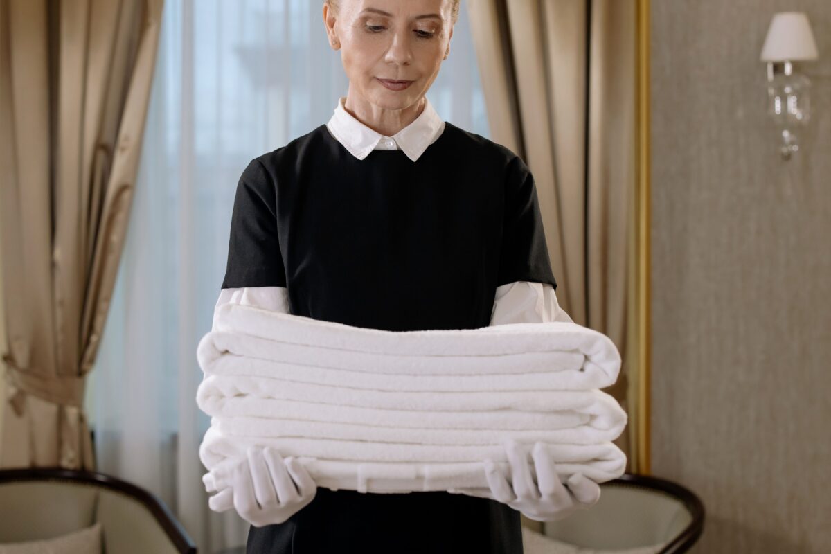 pexels 6466221 | Two Scots Abroad Professional housekeeper holding a stack of fresh towels in a luxurious hotel room, showcasing elegance and service.