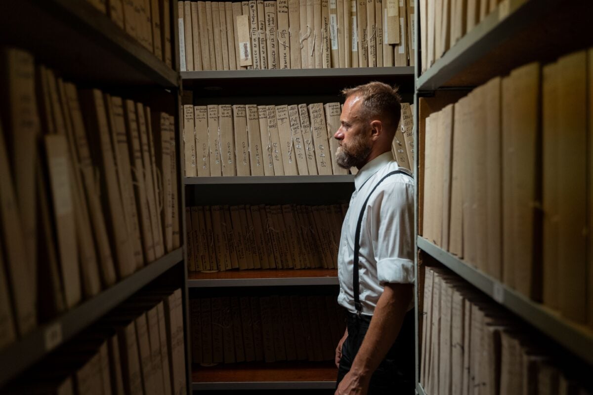 pexels 8872343 | Two Scots Abroad A man in formal attire and suspenders stands in an archive surrounded by documents.