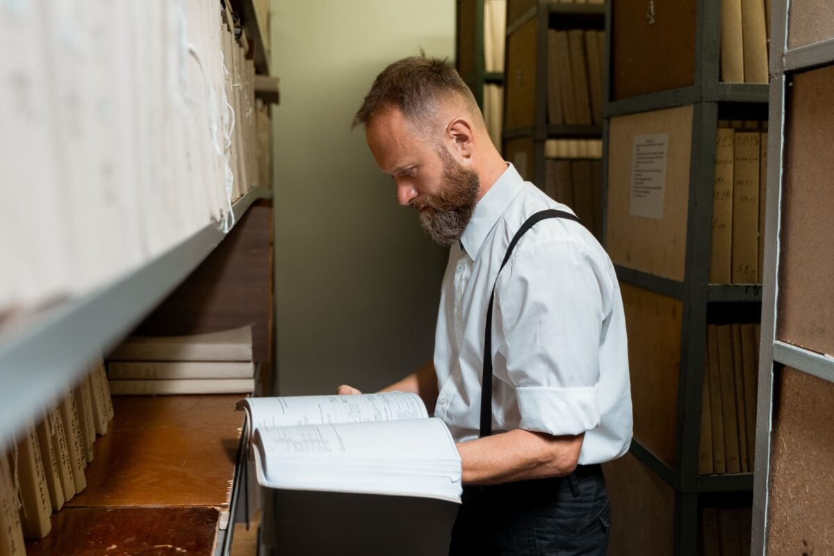 pexels 8872414 | Two Scots Abroad Bearded man in suspenders examining large book in archive room with wooden shelves.