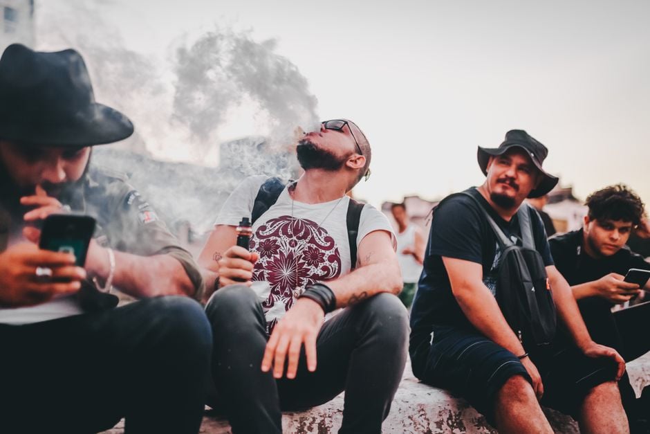 A group of men sitting outdoors, vaping and using mobile phones, in a casual setting.