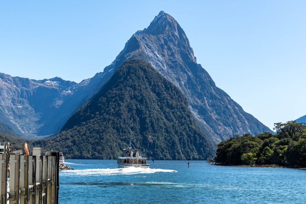 a breathtaking view of milford sound with a boat cruising past majestic mountain | Two Scots Abroad A breathtaking view of Milford Sound with a boat cruising past majestic mountains under a clear blue sky.