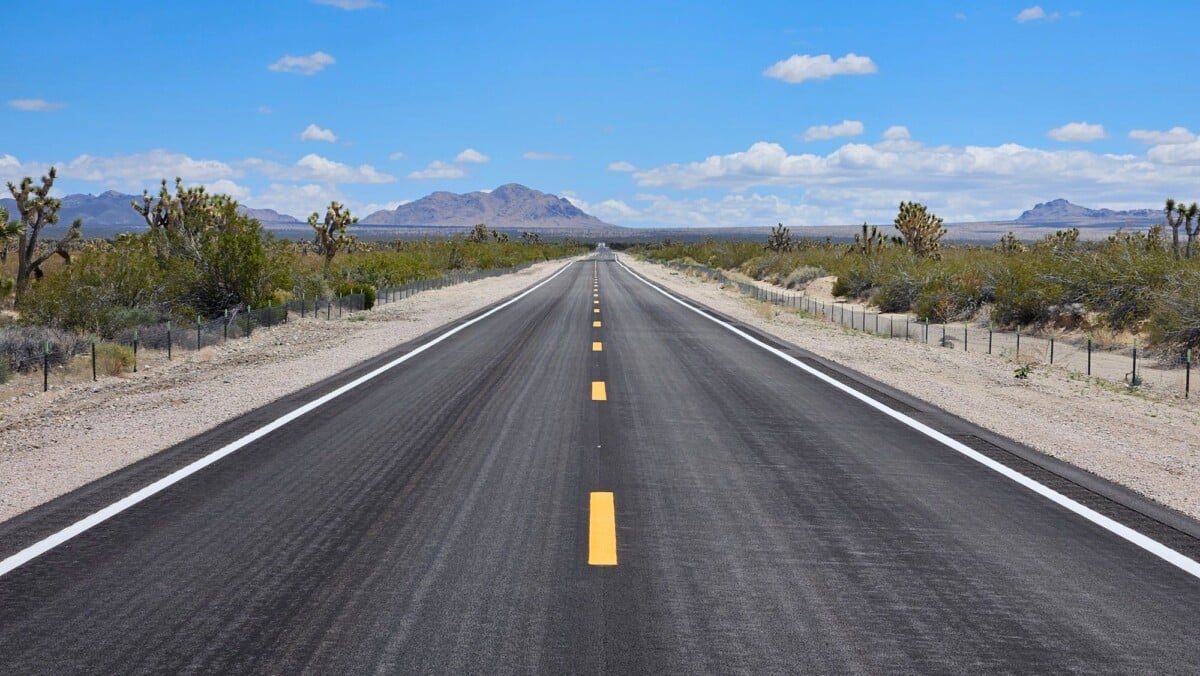 A deserted highway stretches through the Mojave Desert with Joshua trees and mountains under a bright blue sky.