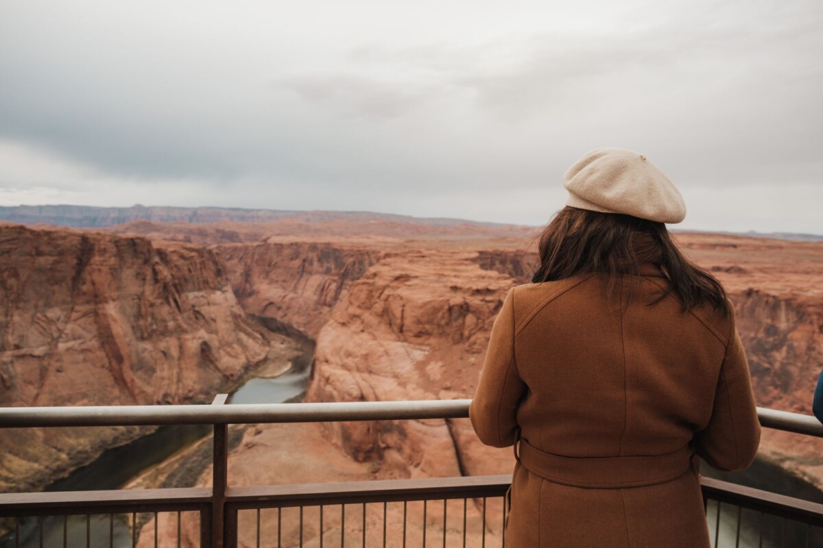 A woman in a coat and beret observes the scenic Horseshoe Bend canyon under an overcast sky.