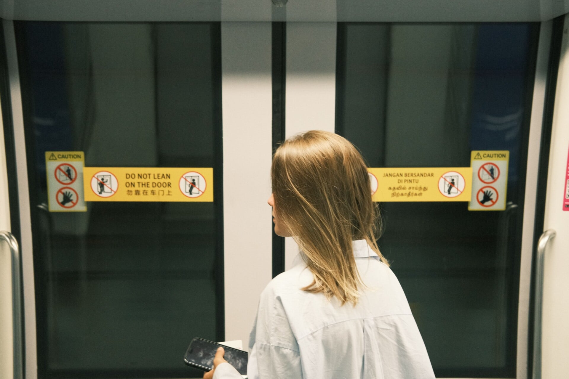 A woman stands in a subway train holding a phone, facing the doors with caution signs.