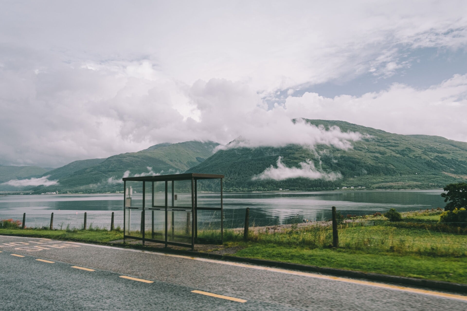 Asphalt roadway near calm water with structure against grassy hilly terrain covered with mist in suburb area on summer day