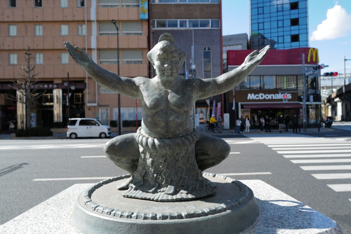 bronze sumo wrestler statue at urban intersection with mcdonalds and modern buil | Two Scots Abroad Bronze sumo wrestler statue at urban intersection with McDonald's and modern buildings in background.