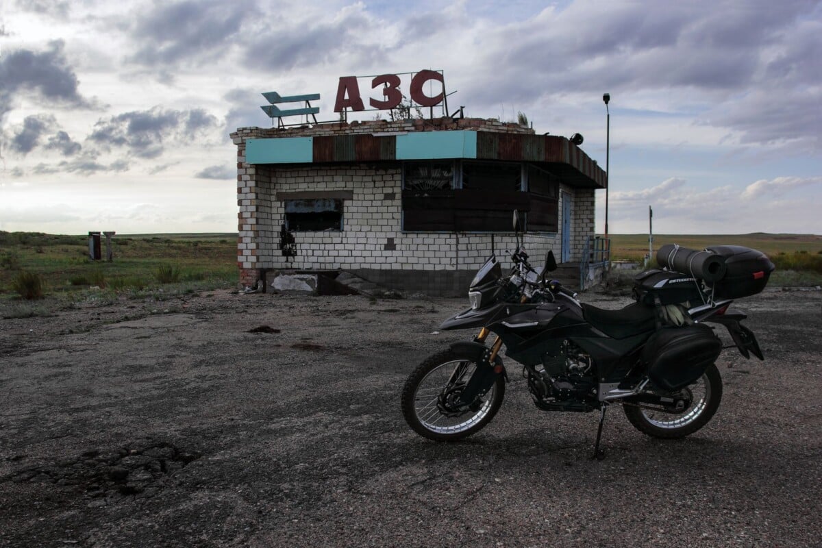 explore adventure with a motorcycle parked at an abandoned rural gas station und | Two Scots Abroad Explore adventure with a motorcycle parked at an abandoned rural gas station under a cloudy sky.