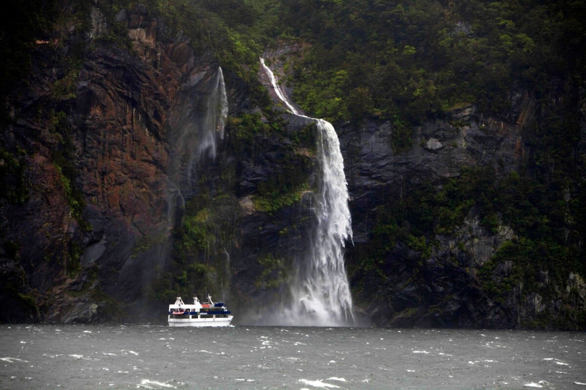 explore the breathtaking view of a majestic waterfall cascading into a fjord wit | Two Scots Abroad Explore the breathtaking view of a majestic waterfall cascading into a fjord with a boat sailing by.