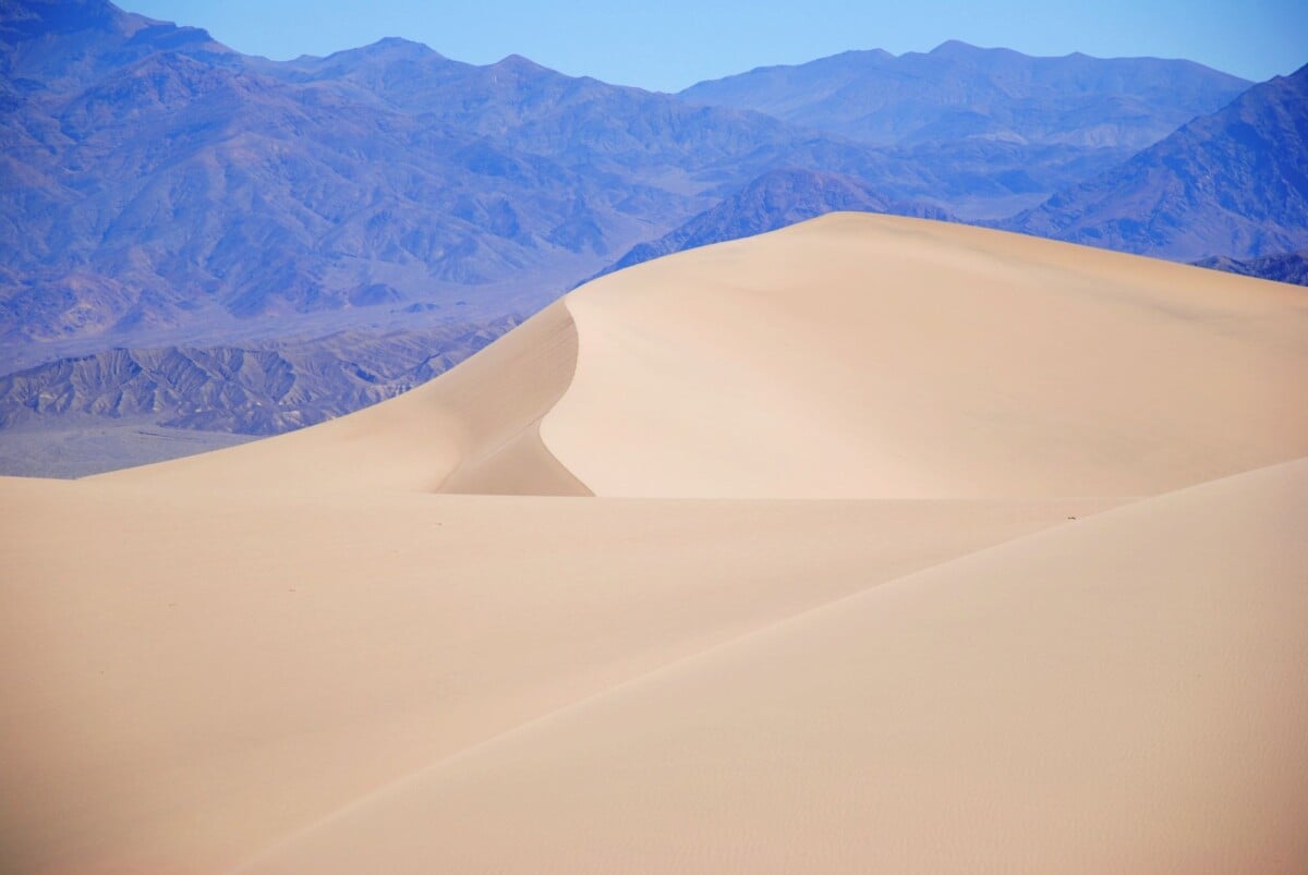 Serene sand dunes in Mojave Desert with distant mountains under clear blue sky.
