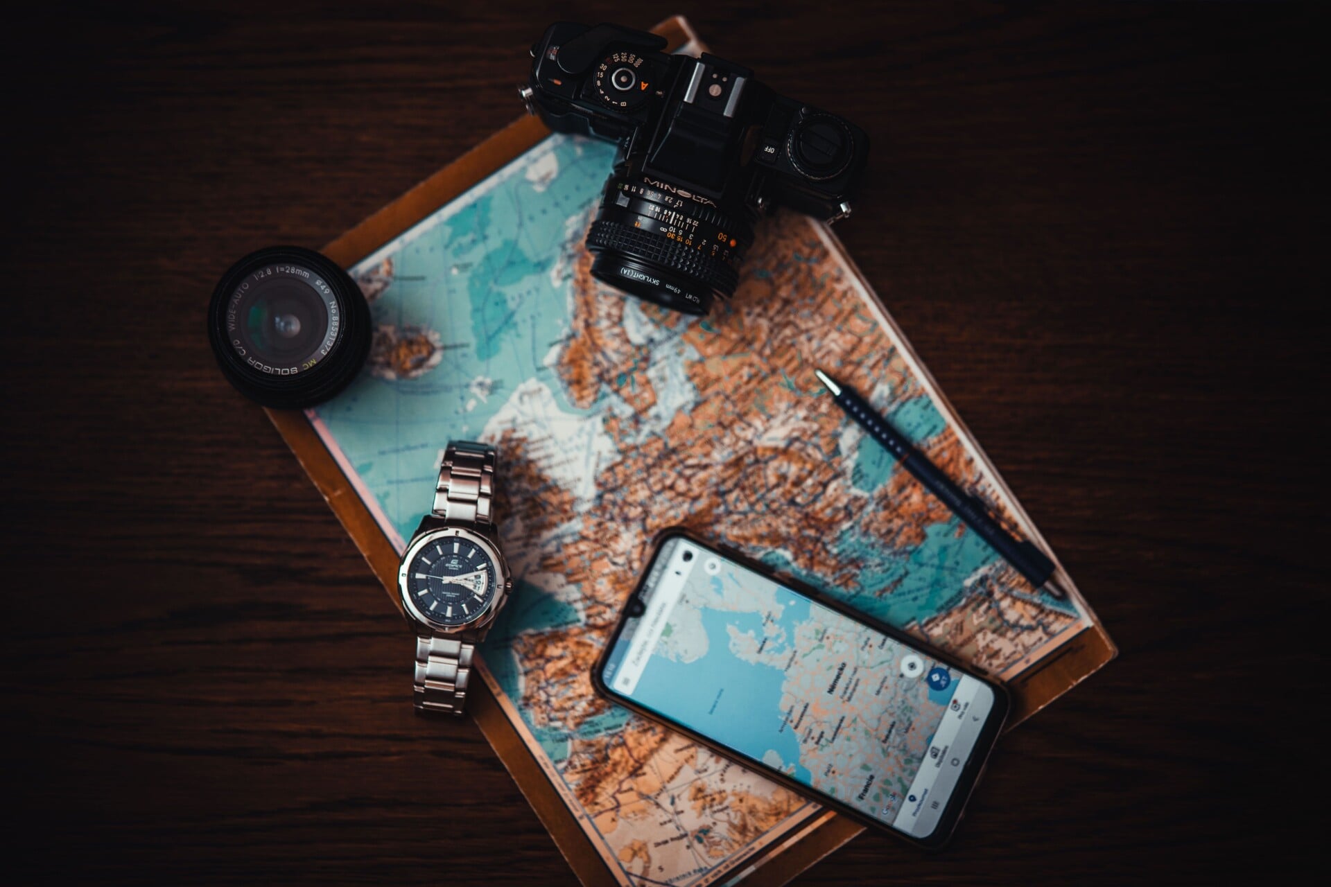 top view of travel essentials including a map camera phone watch and lens on a w | Two Scots Abroad Top view of travel essentials including a map, camera, phone, watch, and lens on a wooden surface.