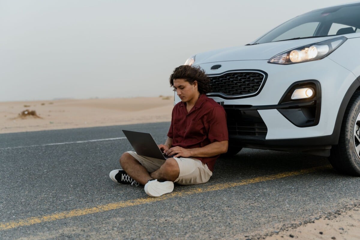 a man using a laptop while sitting on the road beside a parked car in a desert s | Two Scots Abroad A man using a laptop while sitting on the road beside a parked car in a desert setting.
