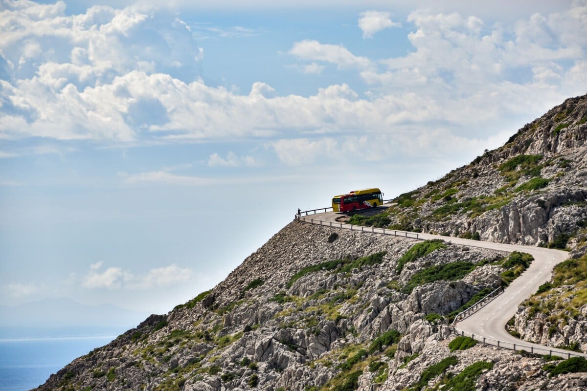 A vibrant bus navigates a narrow, winding road on a rocky mountainside under a cloudy sky.