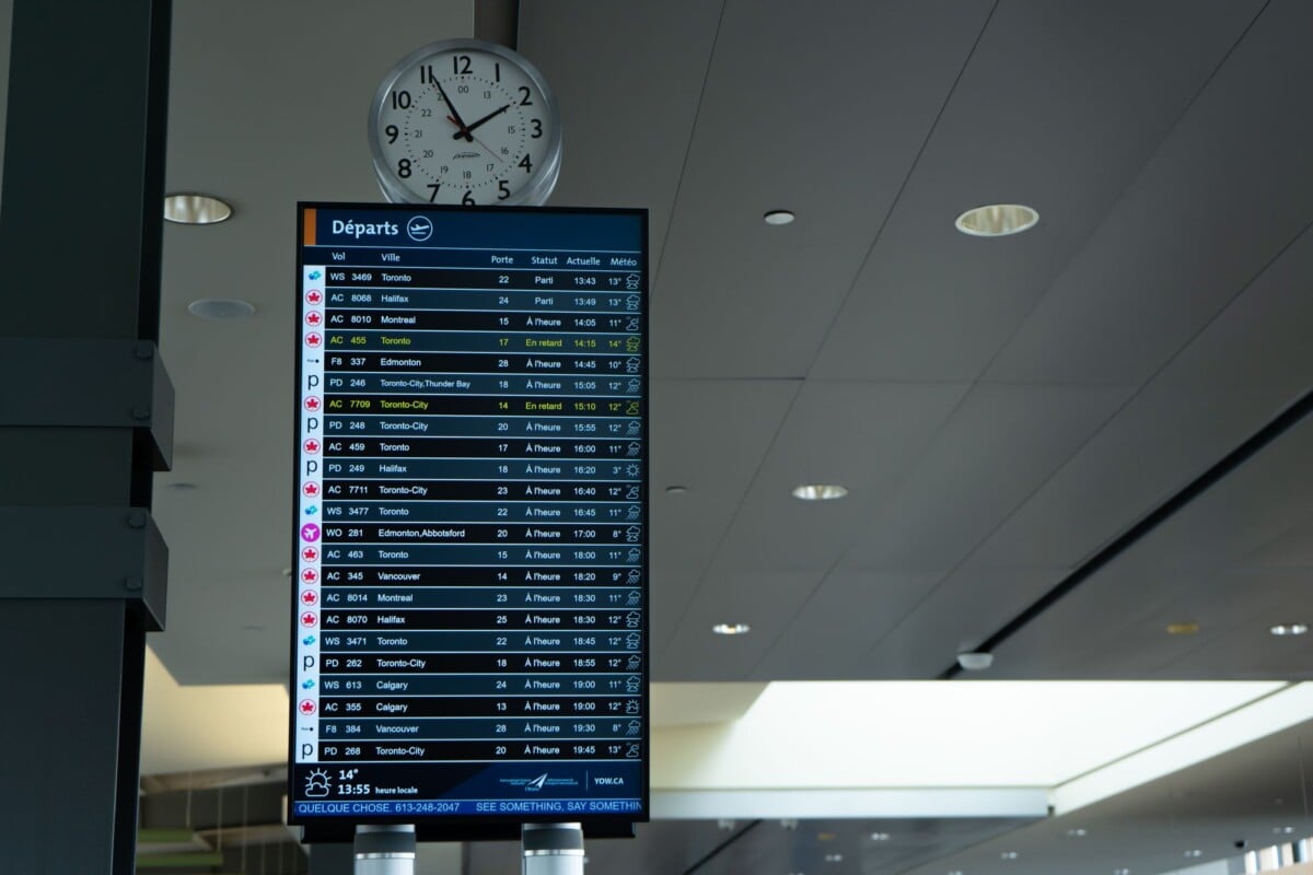 airport departure screen showing various flight schedules and times with a wall | Two Scots Abroad Airport departure screen showing various flight schedules and times with a wall clock above.