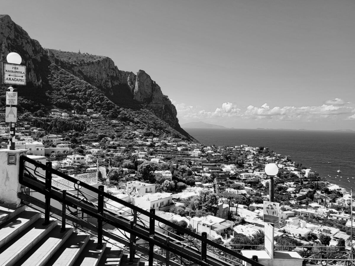 Black and white view of Capri's coastline with townscape and sea under a clear sky.