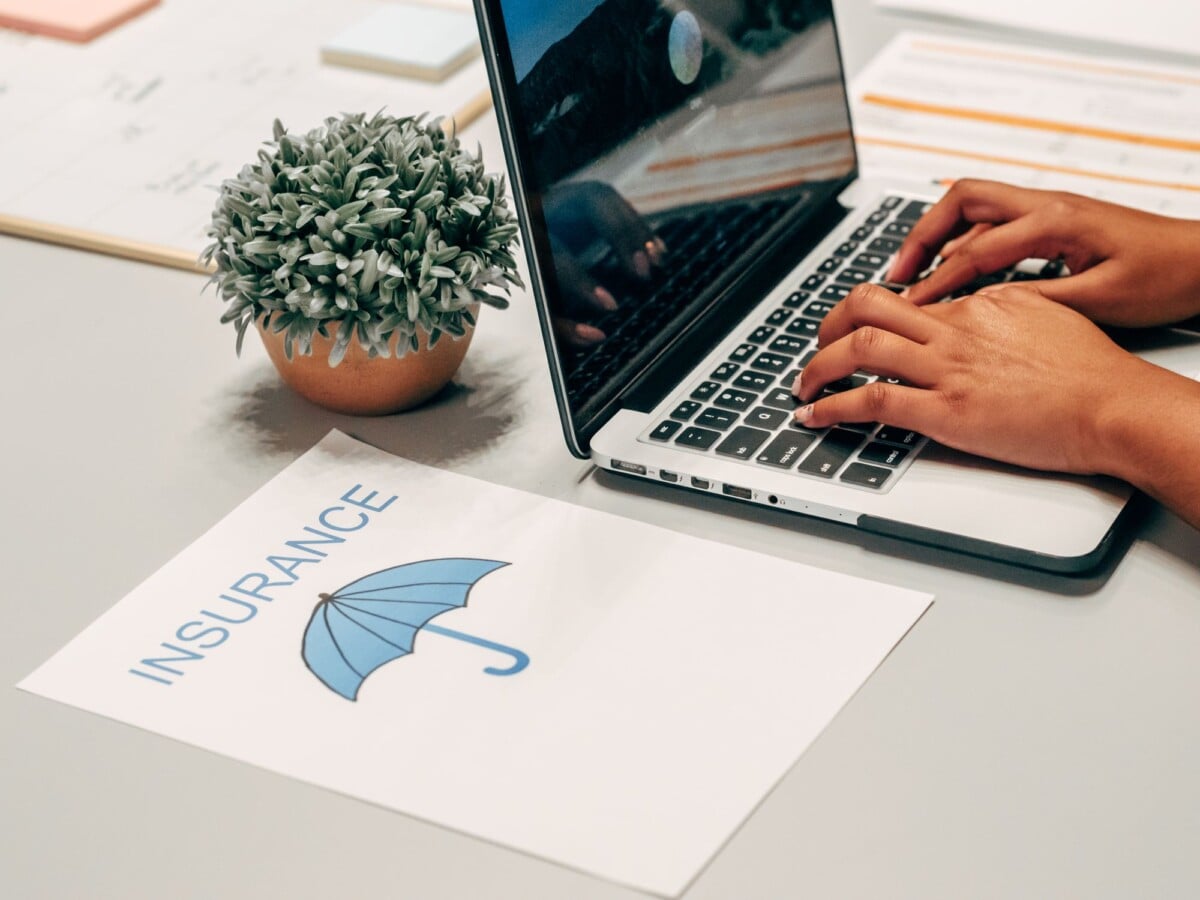 hands typing on a laptop at a desk with an insurance paper and plant suggesting | Two Scots Abroad Hands typing on a laptop at a desk with an insurance paper and plant, suggesting a work environment.