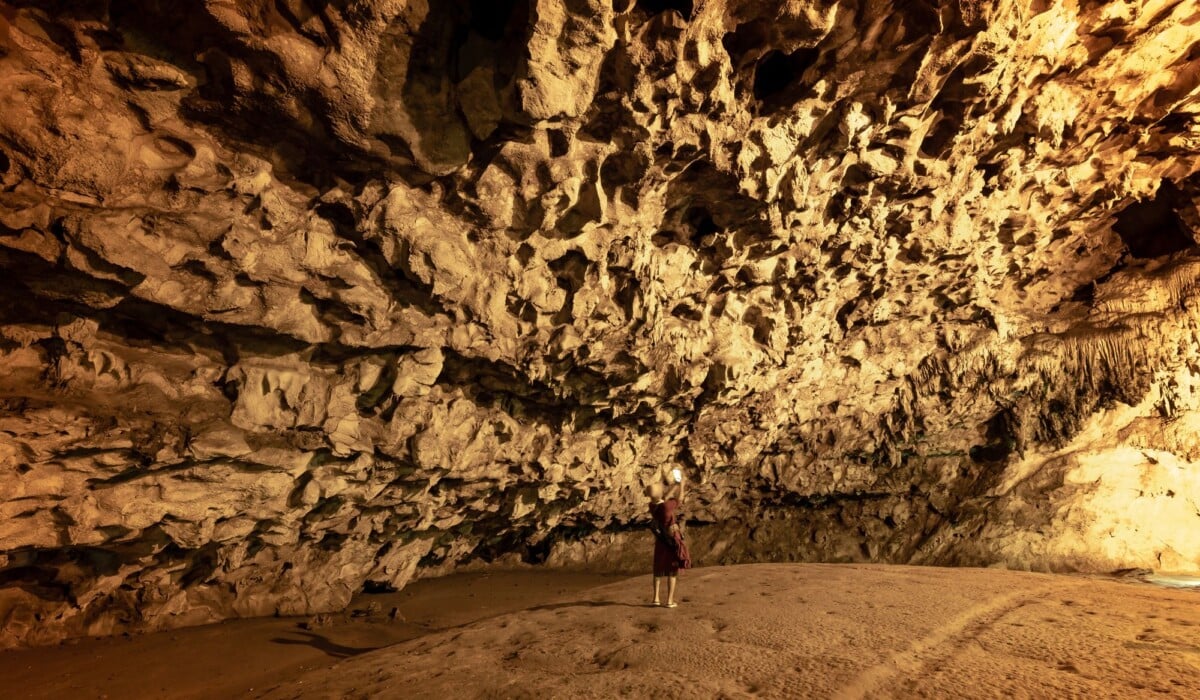 person exploring large cave with limestone formations illuminated by warm light | Two Scots Abroad Person exploring large cave with limestone formations, illuminated by warm light.
