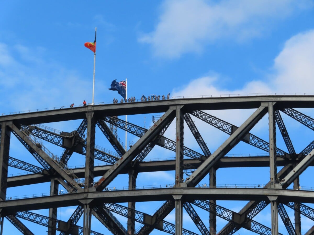 Climbers and an Australian flag on Sydney Harbour Bridge against a blue sky.