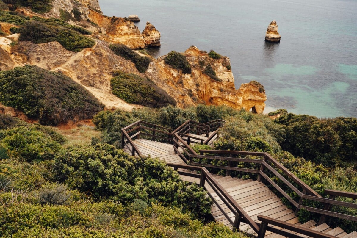 wooden stairs lead to stunning algarve cliffs with lush greenery and turquoise w | Two Scots Abroad Wooden stairs lead to stunning Algarve cliffs with lush greenery and turquoise waters.